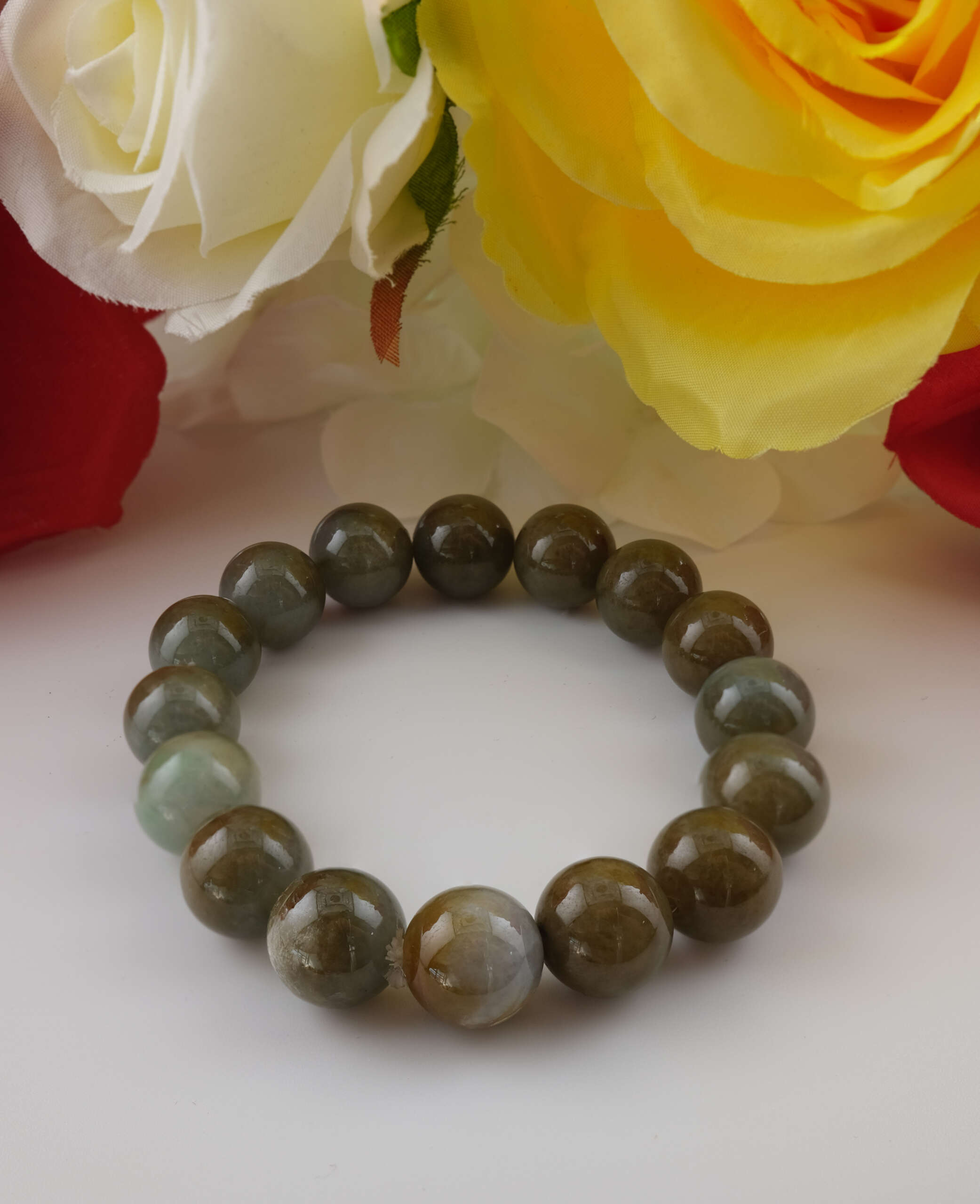 Natural Burmese jadeite bracelet composed of polished 13.4 mm beads, photographed with floral background highlighting smooth luster and earthy green-brown coloration.