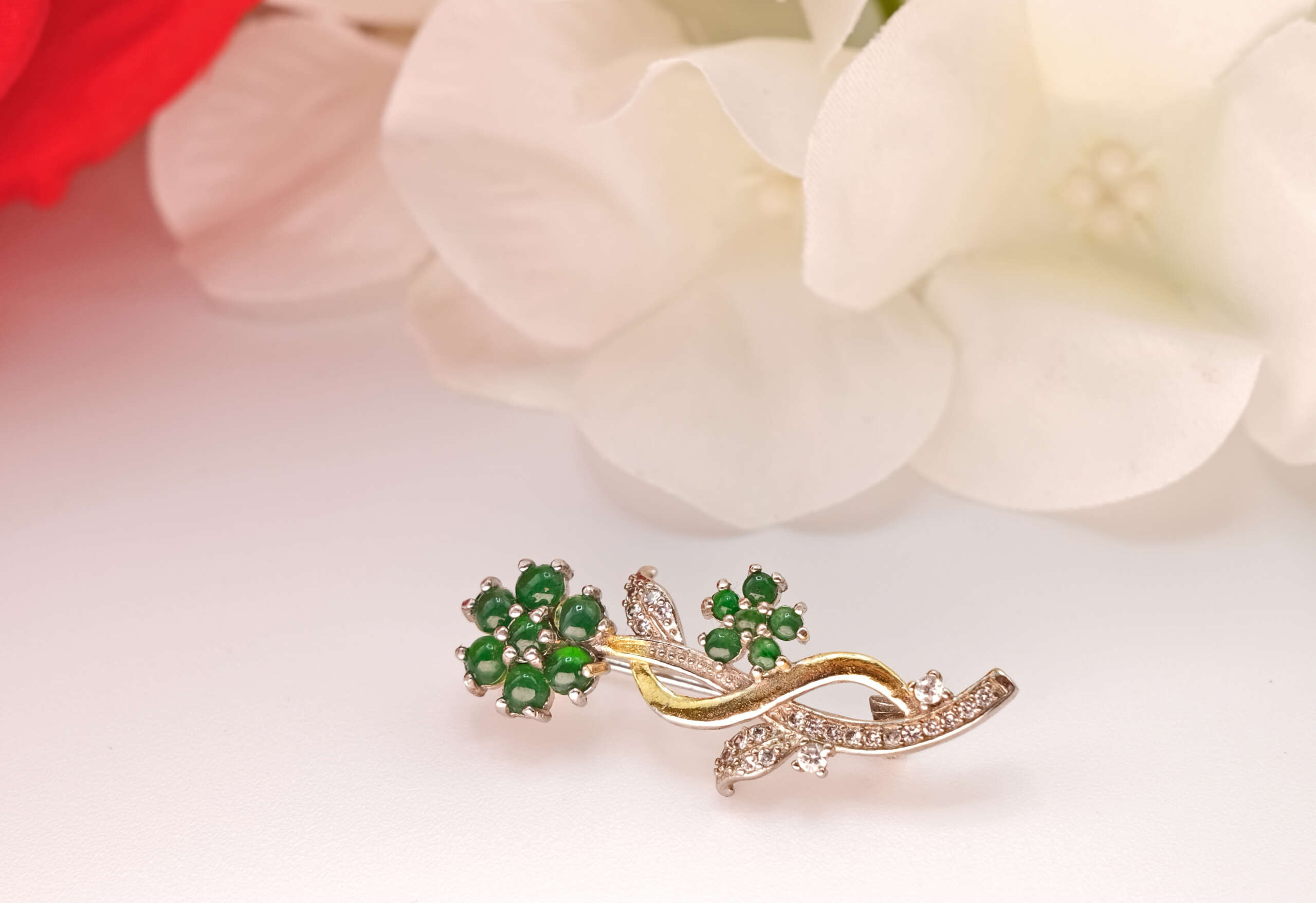 Close-up of natural jadeite flower brooch from Myanmar, set in gold and silver with diamond details, styled against soft white petals to highlight its elegant craftsmanship and vivid green tone.