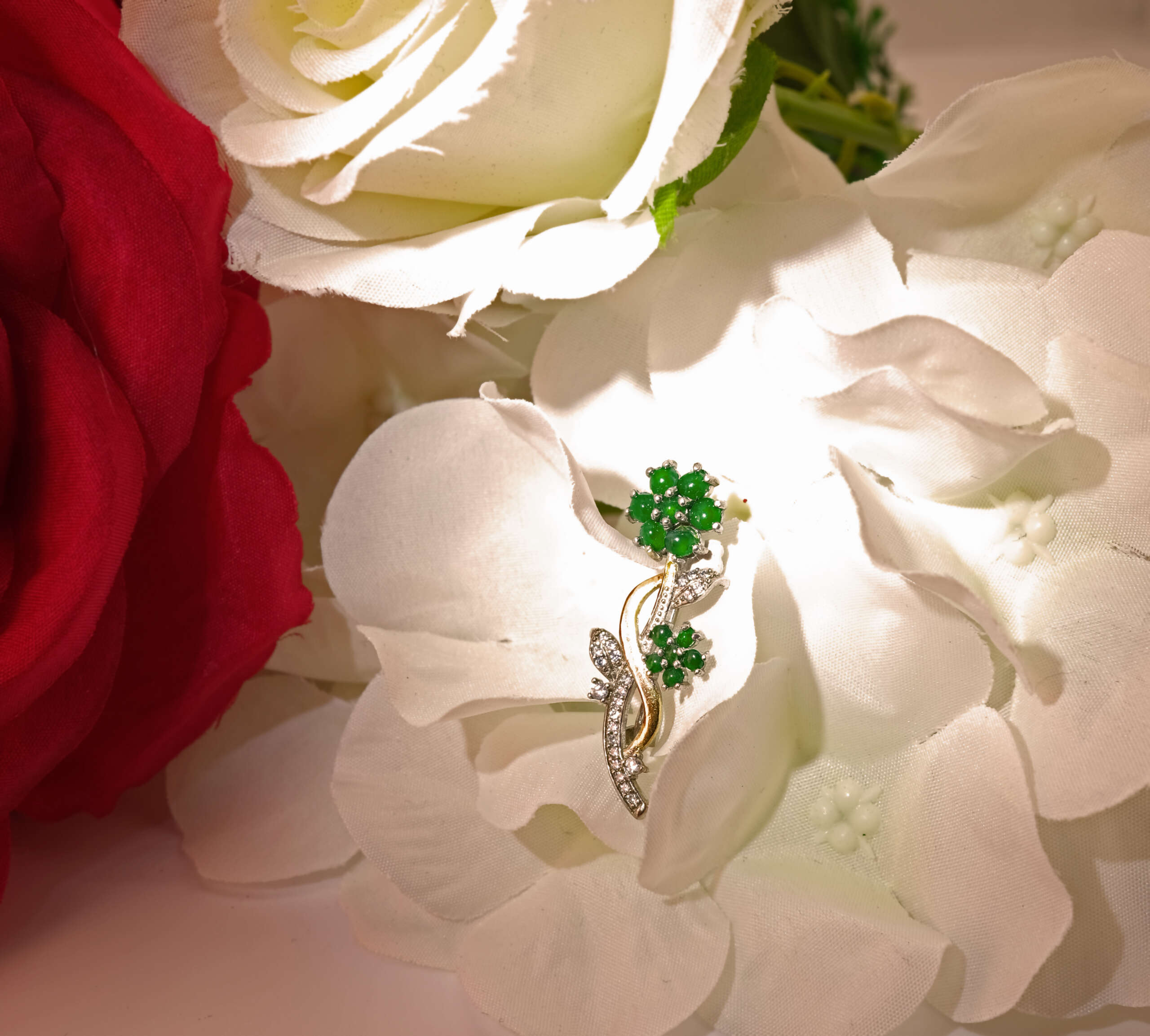 Burmese jadeite floral brooch resting among red and white blossoms under warm light, showcasing Myanmar’s heritage of fine jade craftsmanship in a contemporary NYC jewelry setting.