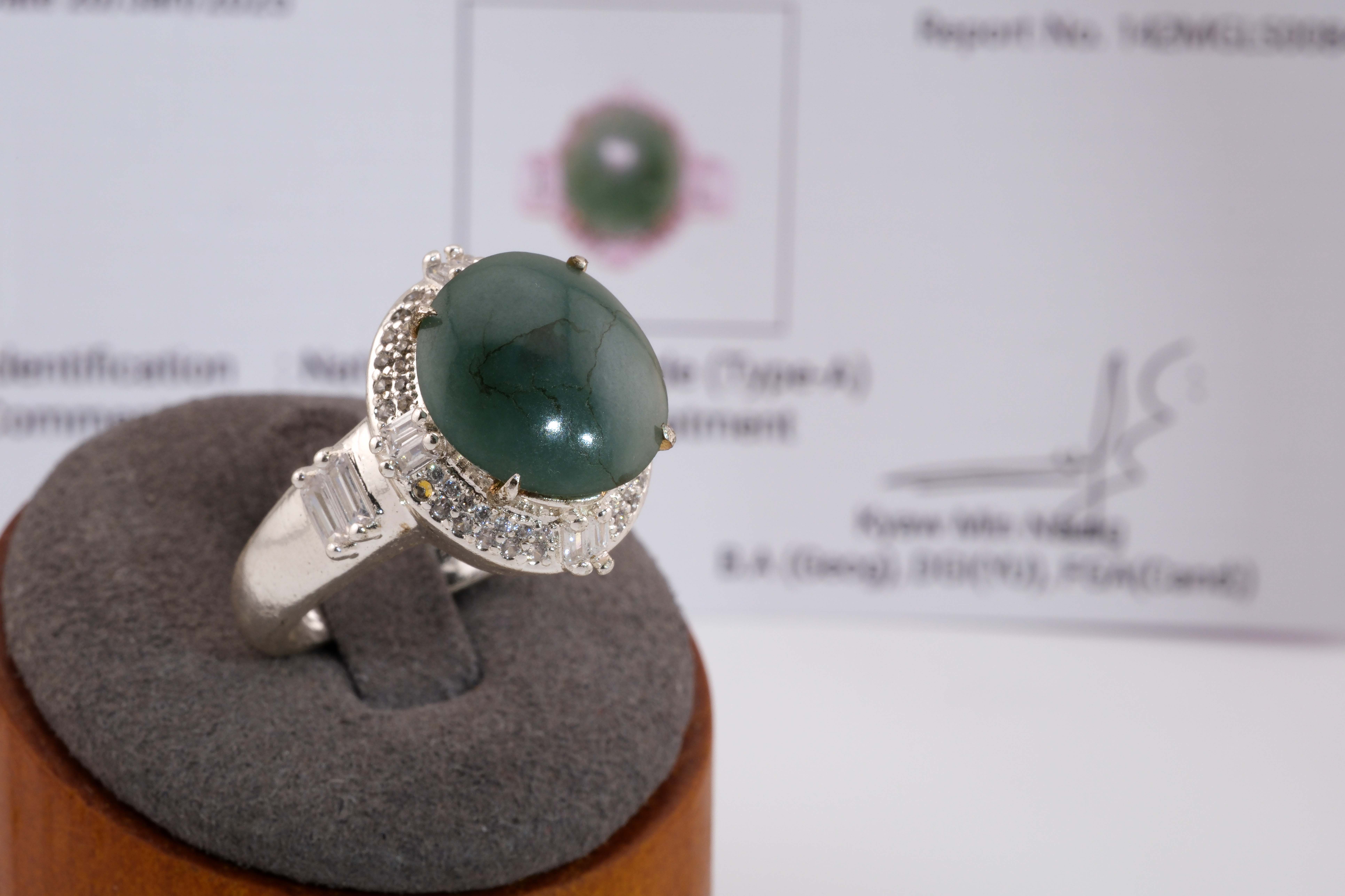 A size 9 natural jadeite jade ring displayed on a gray ring stand with the gemmological certificate blurred behind it. The large bluish-green oval cabochon jade sits in a silver halo setting with baguette and round accent stones