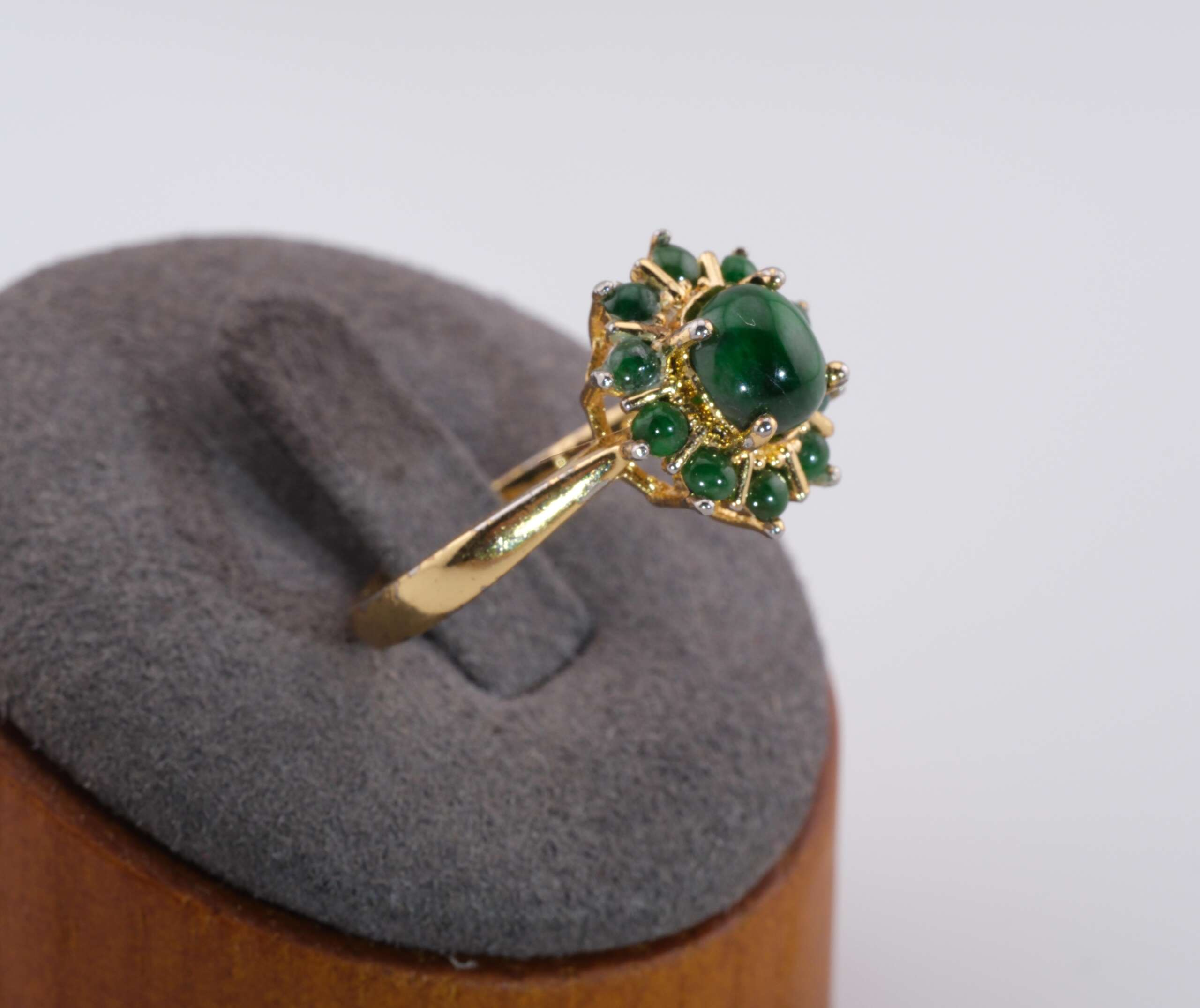 Side-angled view of the gold jadeite cluster ring on a grey display stand, showing the raised floral halo and polished gold tone alloy band.