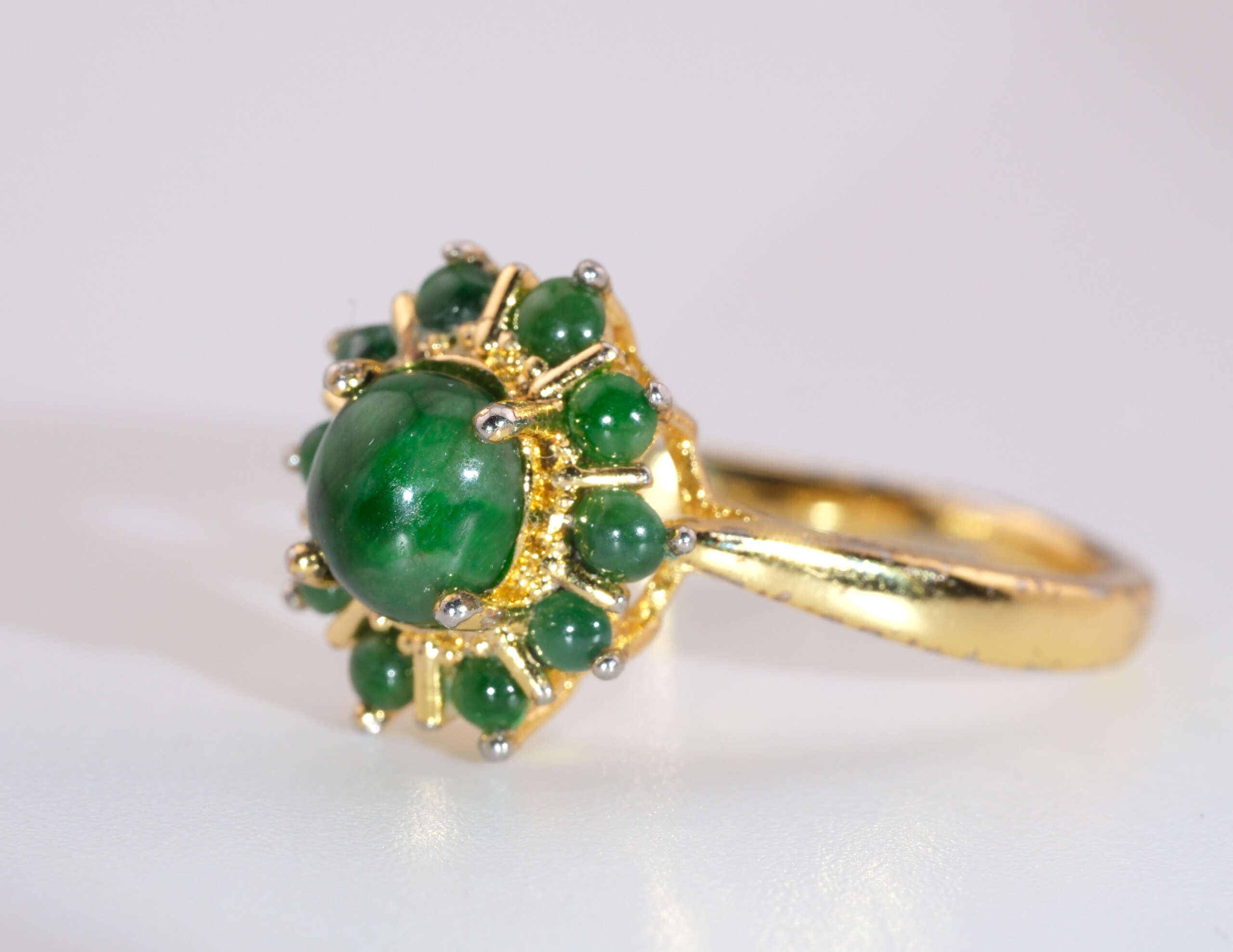 A gold-plated ring featuring a glossy green jadeite cabochon at the center, surrounded by a circular halo of small round jade cabs in a floral arrangement.