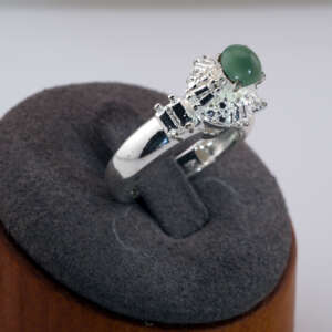 Silver-tone ring with a round green jadeite cabochon displayed on a gray jewelry stand, showing detailed side accents around the stone