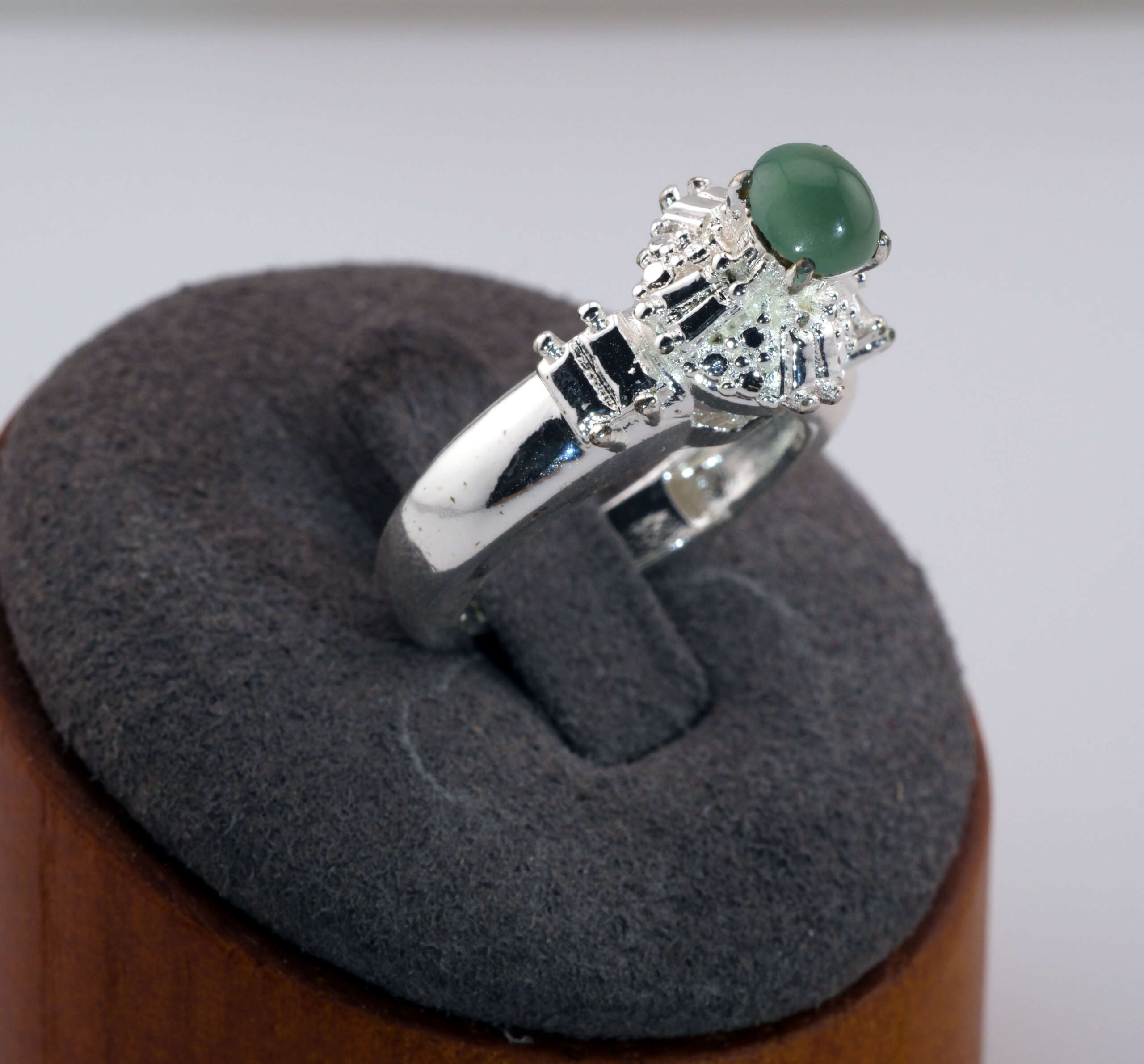 Silver-tone ring with a round green jadeite cabochon displayed on a gray jewelry stand, showing detailed side accents around the stone