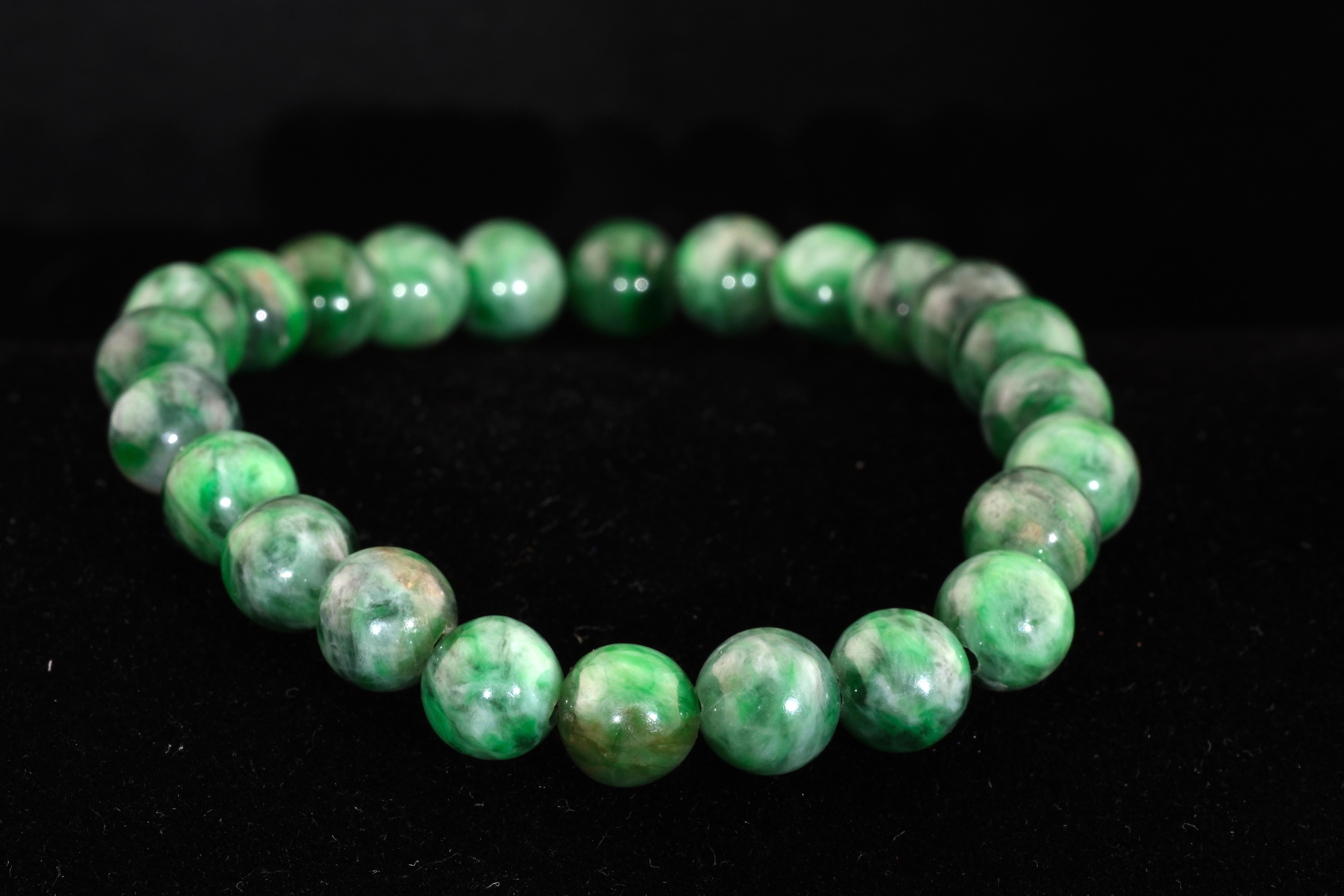 A circular string of polished Maw Sit Sit jadeite beads resting on a black velvet surface, showing vibrant swirling green tones.