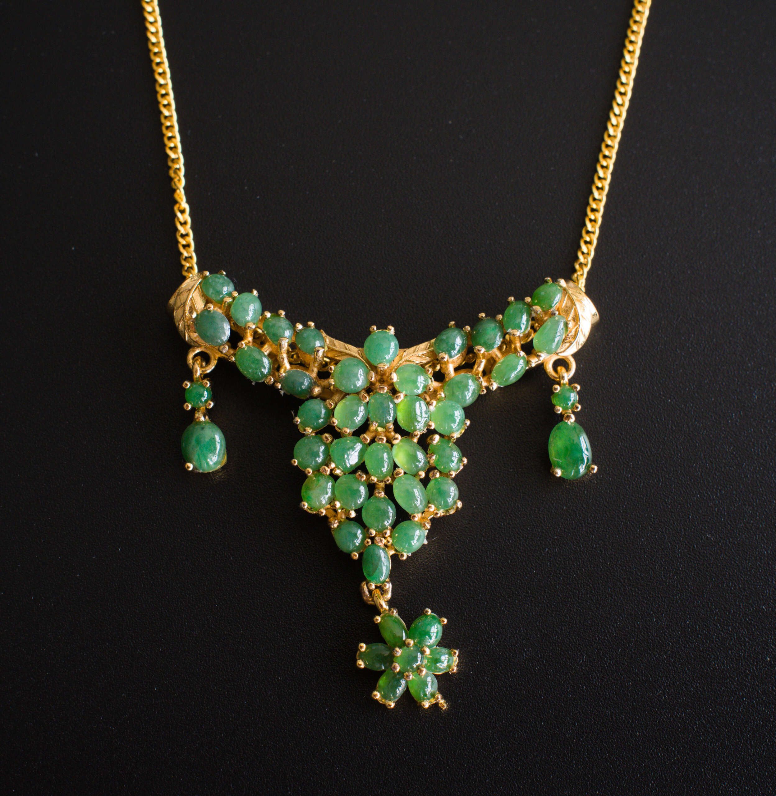 Gold-tone alloy necklace with clustered green jade cabochons, triangular pendant and floral drop, photographed on black background.