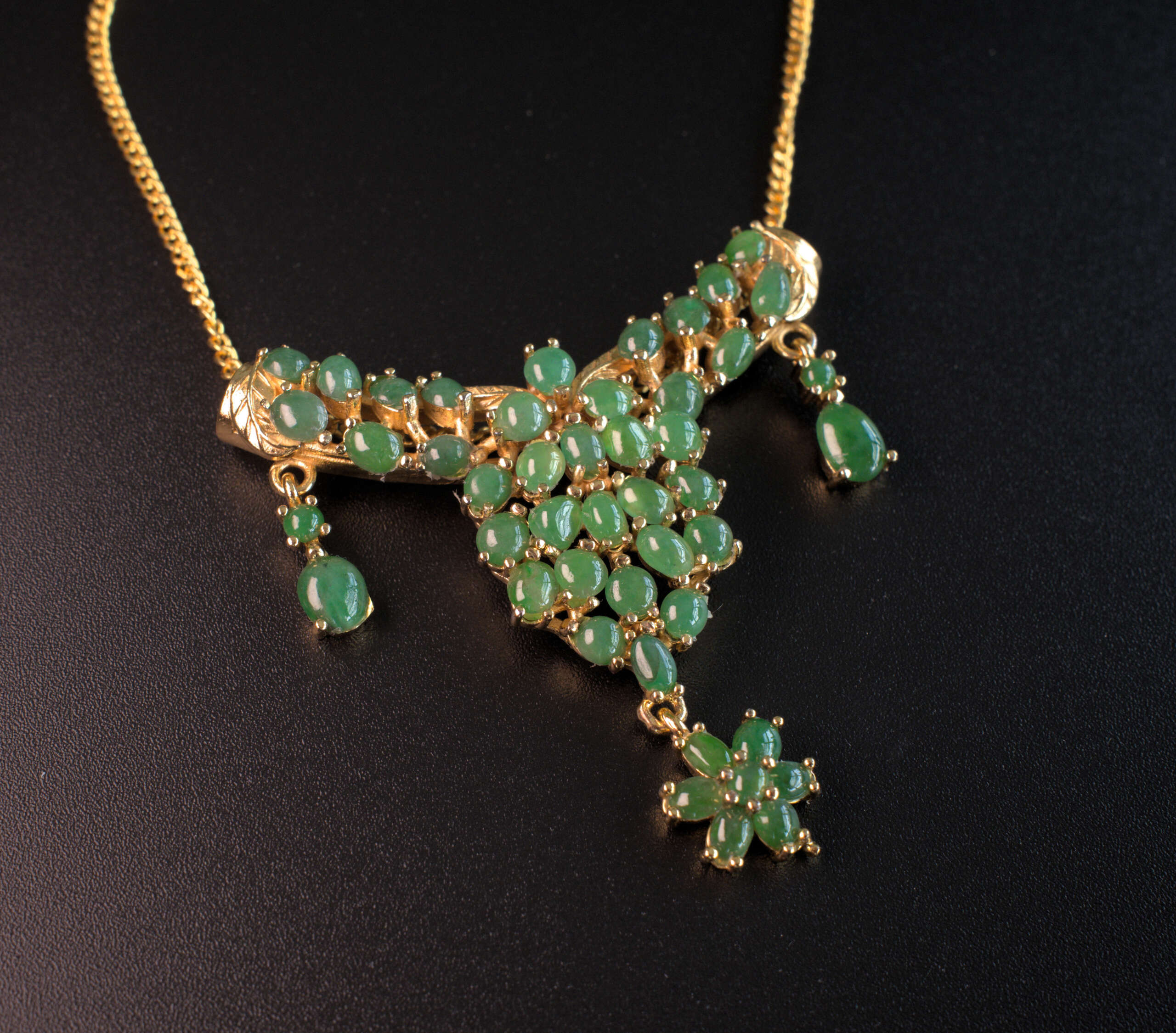 Angled close-up of Myanmar green jade cabochon cluster pendant in gold-tone setting with side dangles and flower drop on black background.