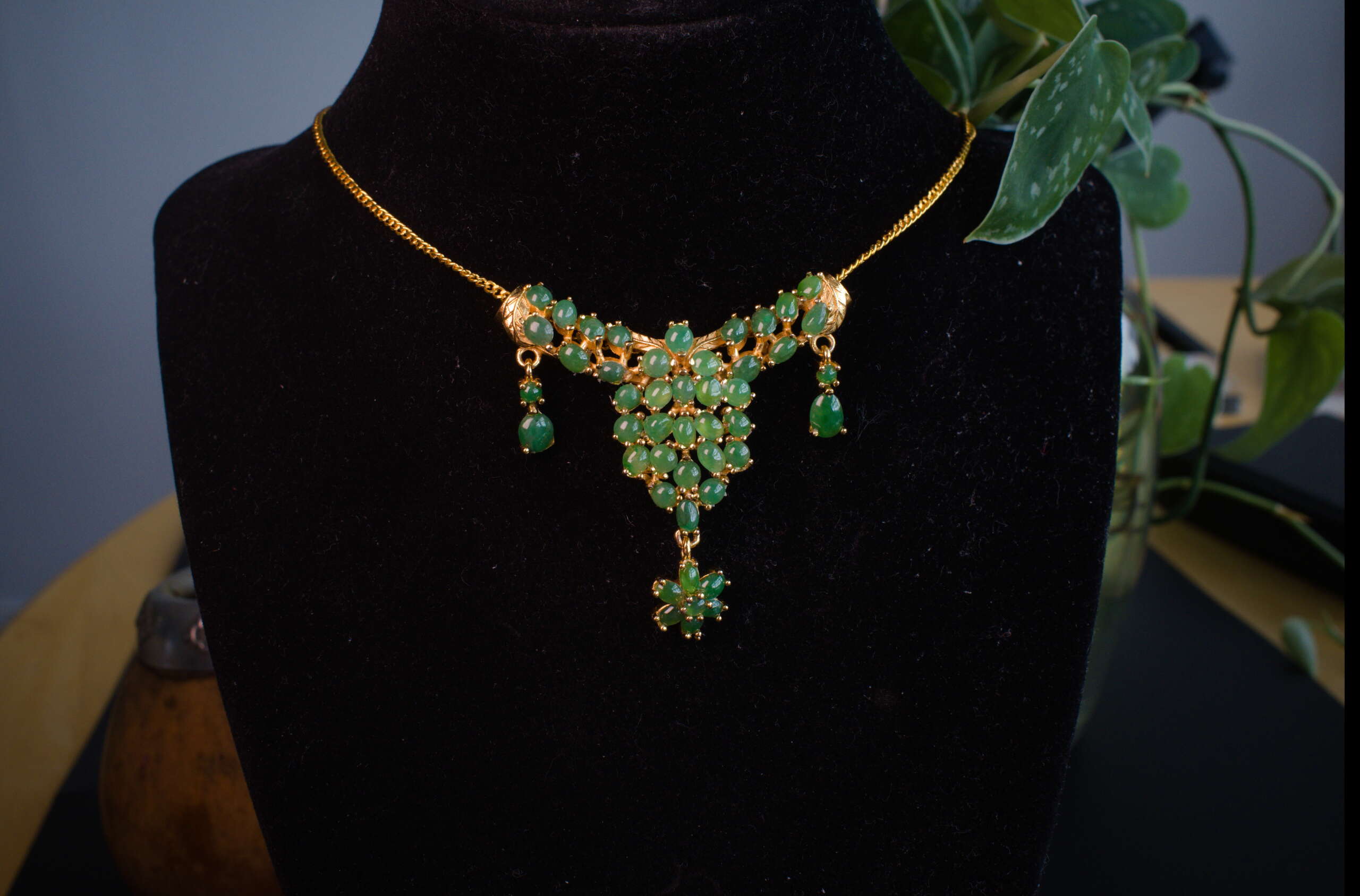 Front view on display bust of gold-tone necklace featuring green jade cabochon cluster pendant with dangling accents and flower-shaped drop.